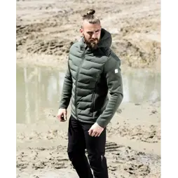 Muž v zelené prošívané bundě s kapucí a černých kalhotách stojí na blátivém povrchu s kalužemi.