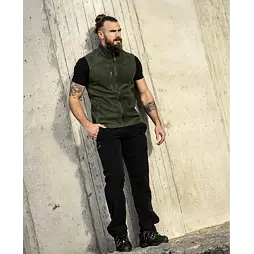 Muž s tetováním na rukou stojí opřený o betonovou stěnu, má svázané vlasy do drdolu a oblečený zelený fleece bez rukávů, černé tričko a černé kalhoty s funkčními prvky.