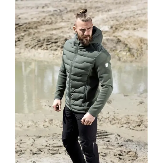 Muž v zelené prošívané bundě s kapucí a černých kalhotách stojí na blátivém povrchu s kalužemi.