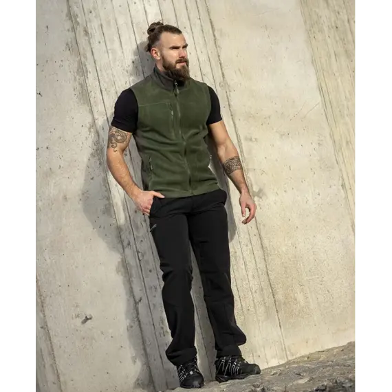 Muž s tetováním na rukou stojí opřený o betonovou stěnu, má svázané vlasy do drdolu a oblečený zelený fleece bez rukávů, černé tričko a černé kalhoty s funkčními prvky.