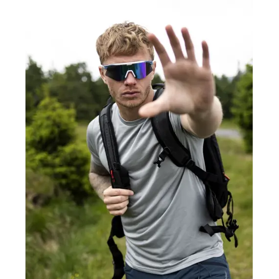 Muž s krátkými vlasy a sportovními brýlemi se zastavuje na turistické stezce v přírodě a natahuje ruku směrem k fotografovi.