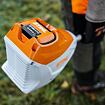 Přenosný akumulátor značky Stihl připevněný k ručnímu elektrickému zahradnímu nářadí držený osobou ve venkovním prostředí.
