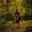 Muž v loveckém oblečení a se zbraní přes rameno kráčí lesní cestou obklopenou stromy a podzimní přírodou.