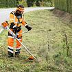 Pracovník v ochranném oděvu se silničním výstražným označením seká trávu a nechtěné rostliny na kraji silnice pomocí motorové křovinořezu.