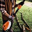 Muž drží elektrický nástroj na zahradní práce značky STIHL, připravený k řezání dvou ležících větví na trávníku.