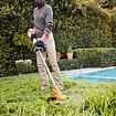 Muž se štítem na obličej a rukavicemi stříhá vysokou trávu kolem bazénu pomocí motorového křovinořezu.