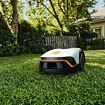 Robotická sekačka trávy pohybující se po zahradě před domem obklopeným stromy a keři.