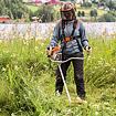 Muž v ochranném oděvu používá motorový křovinořez k sekání vysoké trávy u břehu řeky či jezera.