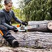 Muž sedí na pařezu a připravuje motorovou pilu ke kácení nebo řezání dřeva v zahradě či lese.