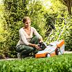 Žena klečí u akumulátorové sekačky na trávu v zahradě a vkládá nebo vyndává baterii.
