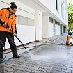 Muž v pracovním oblečení čistí venkovní dlažbu tlakovou myčkou před obytnou budovou.