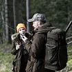 Muž s batohem a zvukovou tlumící návlekem na zbraň komunikuje přes vysílačku, vedle něj žena v oranžové čepici v lesním prostředí.