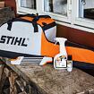 Přepravní taška na motorovou pilu značky STIHL umístěná na dřevěné lavici vedle čisticího spreje a maziva STIHL.