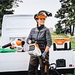 Žena v pracovní ochraně drží zahradní motorový křovinořez značky STIHL před otevřeným nákladním prostorem užitkového vozidla, na kterém jsou další zahradní nástroje a baterie.